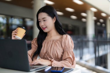 Asyalı bir kadın dizüstü bilgisayarında çalışıyor açık havada içki içiyor genç bir kadın uzaktan çalışmaktan ya da ders çalışmaktan hoşlanıyor.