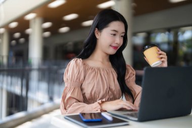 Genç Asyalı kadın dışarıda içkisini yudumlarken taşınabilir bilgisayar kullanıyor. O mutlu görünüyor ve dijital görevleriyle meşgul. Uzak verimlilik kavramları için mükemmel..
