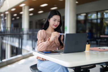 Odaklanmış Asyalı yetişkin bir kadın güneşli bir açık hava ortamında üretken görevler için stilus ile dizüstü bilgisayar kullanıyor ve teknolojinin tadını çıkarıyor.