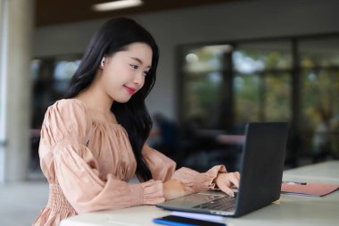 Genç Asyalı kadın dizüstü bilgisayar kablosuz kulaklıklarla meşgul gülümseyen çalışma ortamı modern teknoloji mutlu ruh hali.