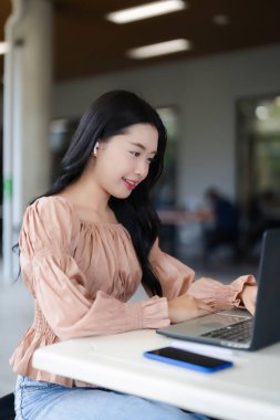 Asyalı genç bir kadın, parlak bir kafede kablosuz kulaklık kullanarak bilgisayarındaki dijital görevlerle uğraşırken gülümsüyor.