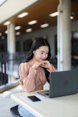 Modern eğitim ortamında veya çalışma alanında kulaklık takan mutlu Asyalı genç kadın dizüstü bilgisayarla meşgul gülümsüyor..