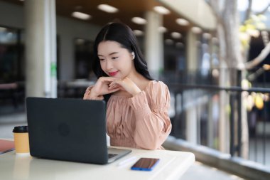 Yetişkin Asyalı kadın dışarıda dizüstü bilgisayara odaklanmış. Uzaktan eğitim, günlük iş görevleri, modern kafe ortamı..