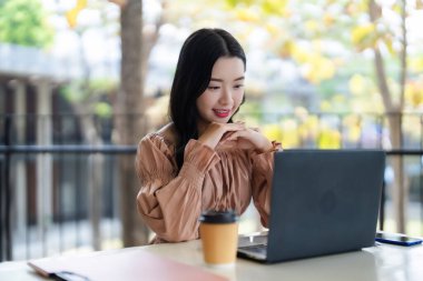 Genç Asyalı yetişkin kadın gülümsemesi online öğrenme ya da açık havada rahat bir ortamda günlük uzaktan çalışma için dizüstü bilgisayar kullanır.