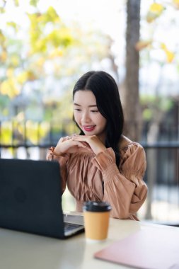 Kahvenin keyfini çıkaran bir kafede dizüstü bilgisayarda çalışan gülümseyen Asyalı kadın. Üretken genç yetişkin kadın çalışmaları veya online iş yönetimi.