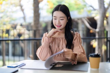 Asyalı yetişkin kadın açık hava belgelerini incelikle inceliyor. Yakınlarda kahve ve akıllı telefon ile odaklanmış bir iş seansından hoşlanıyor..