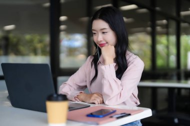 Genç Asyalı kadın, parlak bir ofis ortamında dizüstü bilgisayarına konsantre oluyor. Derin düşüncelere dalmış yeni fikirleri araştırıyor..