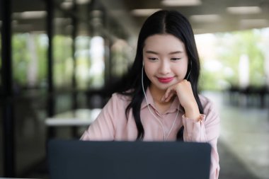 Asyalı yetişkin bir kadın bilgisayarında kulaklık takarak sanal öğrenmeyi seviyor. Çevrimiçi dijital içerikle uğraşan üretken serbest çalışan öğrenci.