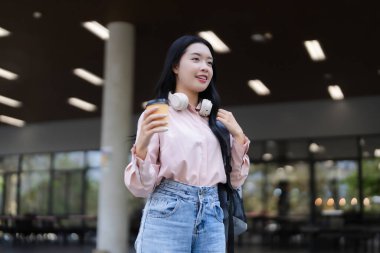 Canlı genç Asyalı kadın ferahlatıcı kahve fincanı tutuyor. Ders için kulaklık takıyor ya da parlak üniversite kampüs binasında çalışıyor.