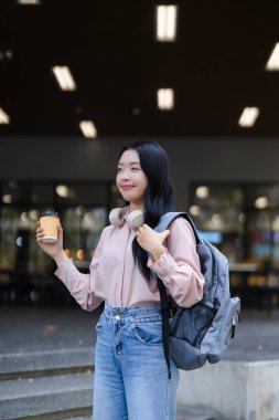 Canlı Asyalı öğrenci modern kampüste sabah kahvesinin tadını çıkarıyor. Genç bir kadın sırt çantası ve kulaklık taşıyor. Kentsel yaşam tarzı.