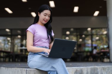 Gülümseyen Asyalı kadın bilgisayarını dışarıda uzaktan çalışmada ya da online çalışmada kullanıyor. Teknolojiyle açık bir ortamda bağlantı kuruyor..