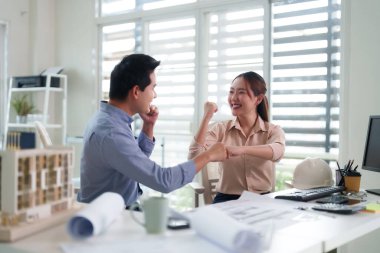 İki gülümseyen Asyalı meslektaşımız projeyi tamamlamak için yumruk tokuşturuyor. Modern ofis ortamında mimari planlar ve başlık mevcut..