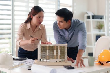 Yetişkin Asyalı mimarlar ve mühendisler, modern bir ofiste detaylı bir mimari modeli inceleyen karmaşık bina tasarımı planlarını işbirliği içinde tartışırlar.