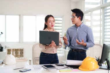 Yetişkin Asyalı profesyoneller planları incelemek için işbirliği yapıyor. Mimar ve mühendis takımı dizüstü bilgisayarla mimari modeli inceliyor. Dinamik ofis toplantı yeri etkileşimi.