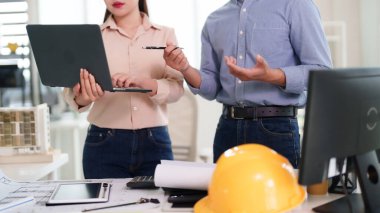 Yetişkin Asyalı mimarlar bir ofis masasında bina planlarını gözden geçiriyorlar. Onlar bir geliştirme projesi tasarımı üzerinde işbirliği yapıyorlar. Teknoloji araçları çalışmalarını geliştirir.