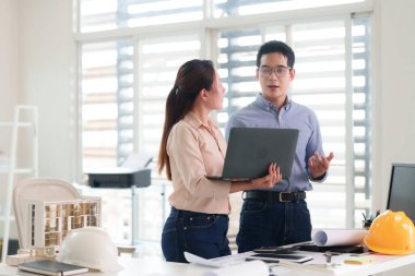 Asyalı yetişkinler modern ofis ortamında mimari tasarım projesinde işbirliği yapıyorlar. Teknoloji ve takım çalışması kullanarak planları tartışıyorlar.