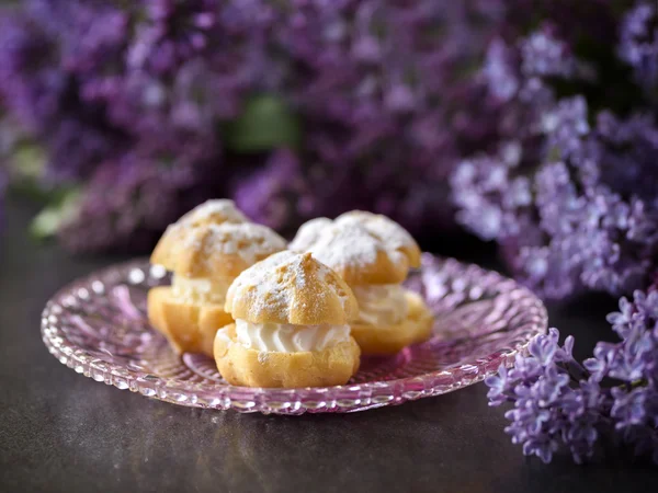 Crema de hojaldre o profiterol con relleno, cobertura de azúcar en polvo y flores lila aisladas, sobre fondo oscuro — Foto de Stock