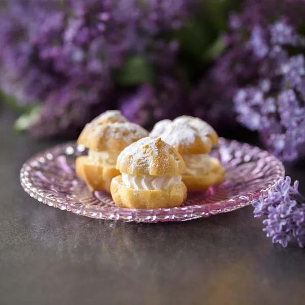 Cream puff or profiterole with filling, powdered sugar topping and lilac flowers isolated, on dark background — Stock Photo, Image