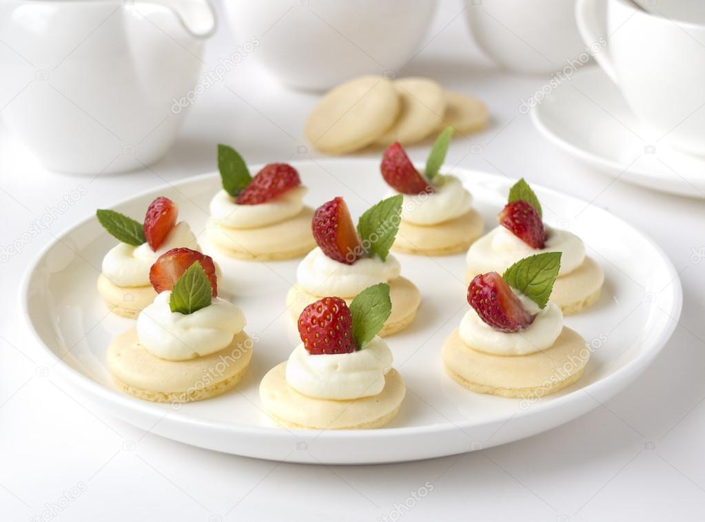 Collection of small cakes with whipped cream, fruits, mint on white plate on table against background