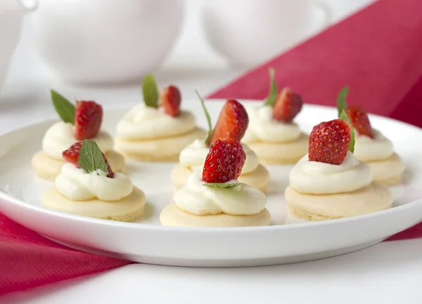 Små kakor med vispad grädde, strawsberry, mint på vit platta på bordet mot ljus bakgrund — Stockfoto