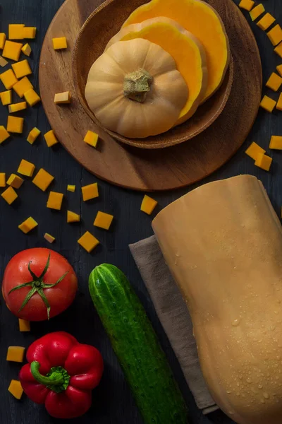 The vertical photo shows a picture of food on a black wooden background. This is a bottle-shaped pumpkin, red tomato, sweet bell pepper, cucumber. Nearby you can see a kitchen towel, wooden plates.