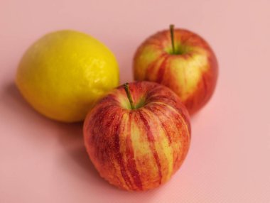 İki olgun gala elması ve bir limon pembe arka planda yatar.
