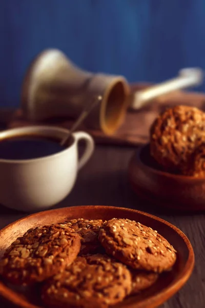 The photo shows an image of food and related objects. There are oatmeal cookies with cereals, a black candle, a white coffee cup, a small spoon. Also wooden kitchen items