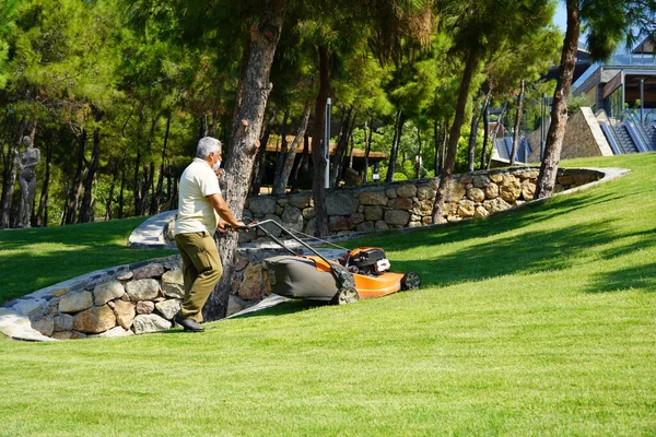 Bodrum, Turquie - août, 2020 Homme adulte en chapeau et masque coupe pelouse dans la journée chaude d'été. Travailler dans la maison et le jardin, prendre soin du territoire. Personnel de service à l'hôtel Photo De Stock