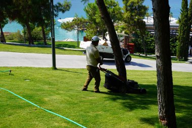 Bodrum, Türkiye - 2020 Ağustos, sıcak yaz günlerinde şapka ve maske takan yetişkin adam çimenleri kesiyor. Evde ve bahçede çalış, toprakla ilgilen. Oteldeki servis elemanları. 
