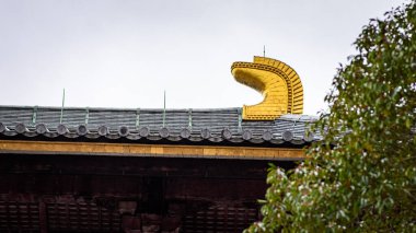 A close-up view of a traditional Japanese temple roof with a distinctive golden ornament shaped like a wave, surrounded by greenery
