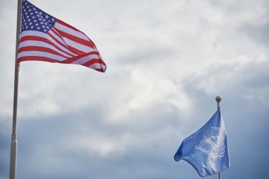 Amerikan ve Birleşmiş Milletler bayrakları bulutlu bir gökyüzüne doğru dalgalanıyor.