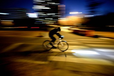 Bir bisikletçinin silüeti, arka planda bulanık ışıklar ve hareket ve hız hissi yaratarak, gece bir şehir caddesinde hızla ilerliyor.