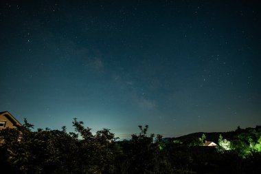 Ağaçlar ve evlerle dolu kırsal bir arazide yıldızlı gece gökyüzü.