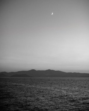 Uzak dağlarla dolu sakin bir denizin üzerinde hilal şeklindeki bir ayın siyah beyaz fotoğrafı..