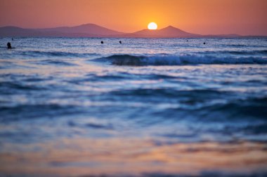 Uzak dağlar ve yüzücülerle sakin bir okyanusun üzerinde gün batımı.