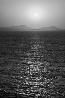 Uzak tepeleri ve batan güneşi olan sakin bir okyanusun siyah ve beyaz fotoğrafı..
