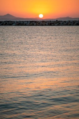 Sakin bir denizin üzerinde sakin bir günbatımı, önü kayalık bir kıyı şeridi ve ufukta uzak dağlar