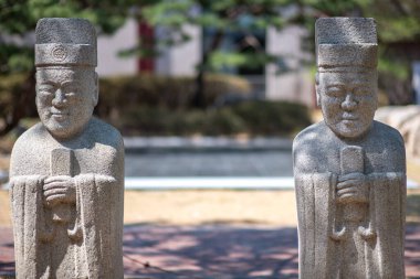 Geleneksel giysiler içinde, arka planda bulanık ağaçlar ve patika olan bir parkta duran iki eski Koreli bilim adamının heykeli..