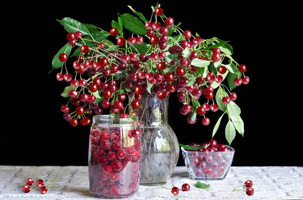 Cold drink with cherry berries in a jar, in a vase and a bouquet of ripe cherries on the table, and a black background