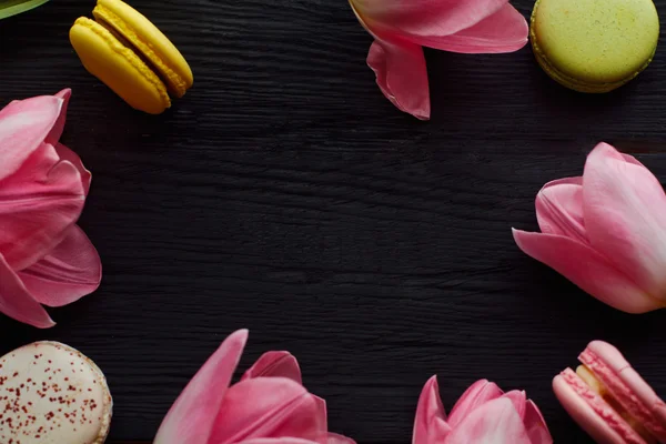 A beautiful bouquet of pink tulips with colorful macaroons laid out as a frame with a free center on dark wooden background — Stock Photo, Image