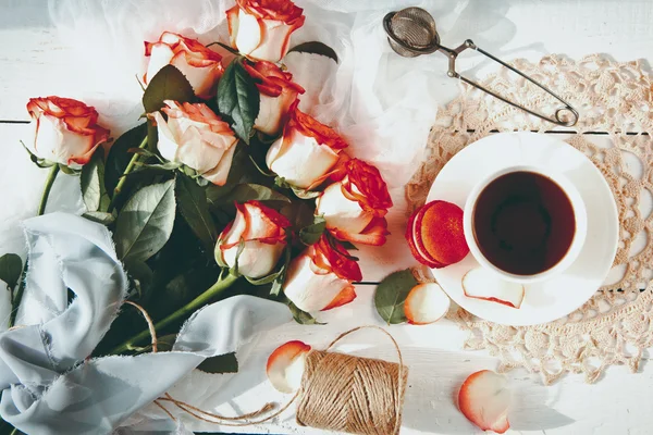 Een mooi boeket rozen, tea Cup, met bitterkoekjes op noodlijdende witte houten tafel. vintage stijl — Stockfoto