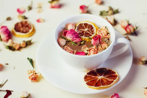 Kräutertee in weißer Tasse und Untertasse mit Rosen und getrockneten Blumen getrocknete runde Zitronenscheiben auf weißem Hintergrund — Stockfoto