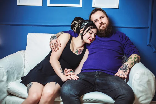 Young couple in love with tattoos hugging and holding hands sitting on sofa — Stock Photo, Image