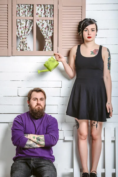 A guy and a girl with tattoos having fun, a man sits on the bench and the girl standing with a watering can — Stock Photo, Image