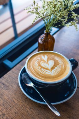 Latte kahve sanatının ahşap masadaki siyah bardaktaki klasik ton görüntüsü. (dikey)