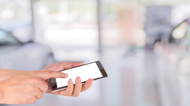 Erkek eli modern bir dokunmatik ekranlı telefon tutuyor ve ticari arabaların görüntüsü araba dükkanının vitrininde duruyor..