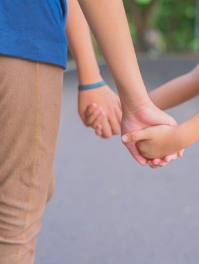 Kız ve erkek kardeşlerin parkta yürürken el ele tutuşmaları arka planda yeşil bir ağaç ile..