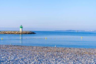 Denizin arka planında deniz feneri ve küçük taşlardan oluşan plajda deniz tamamen sakin ve berrak. Mavi gökyüzü. 