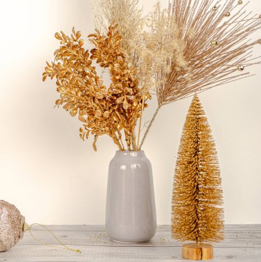  Beyaz bir duvar ve ahşap masa arkaplanındaki vazoda, yeni yıl altın dallı otların kompozisyonu. Altın fidan ağacı vazonun yanında. Tatil dekorasyonu kavramı