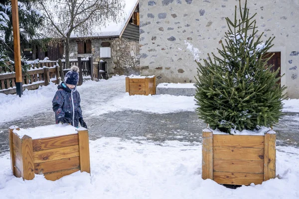Un niño jugando cerca de un pequeño abeto nevado en una maceta de madera al aire libre en invierno. Concepto de Navidad — Foto de Stock